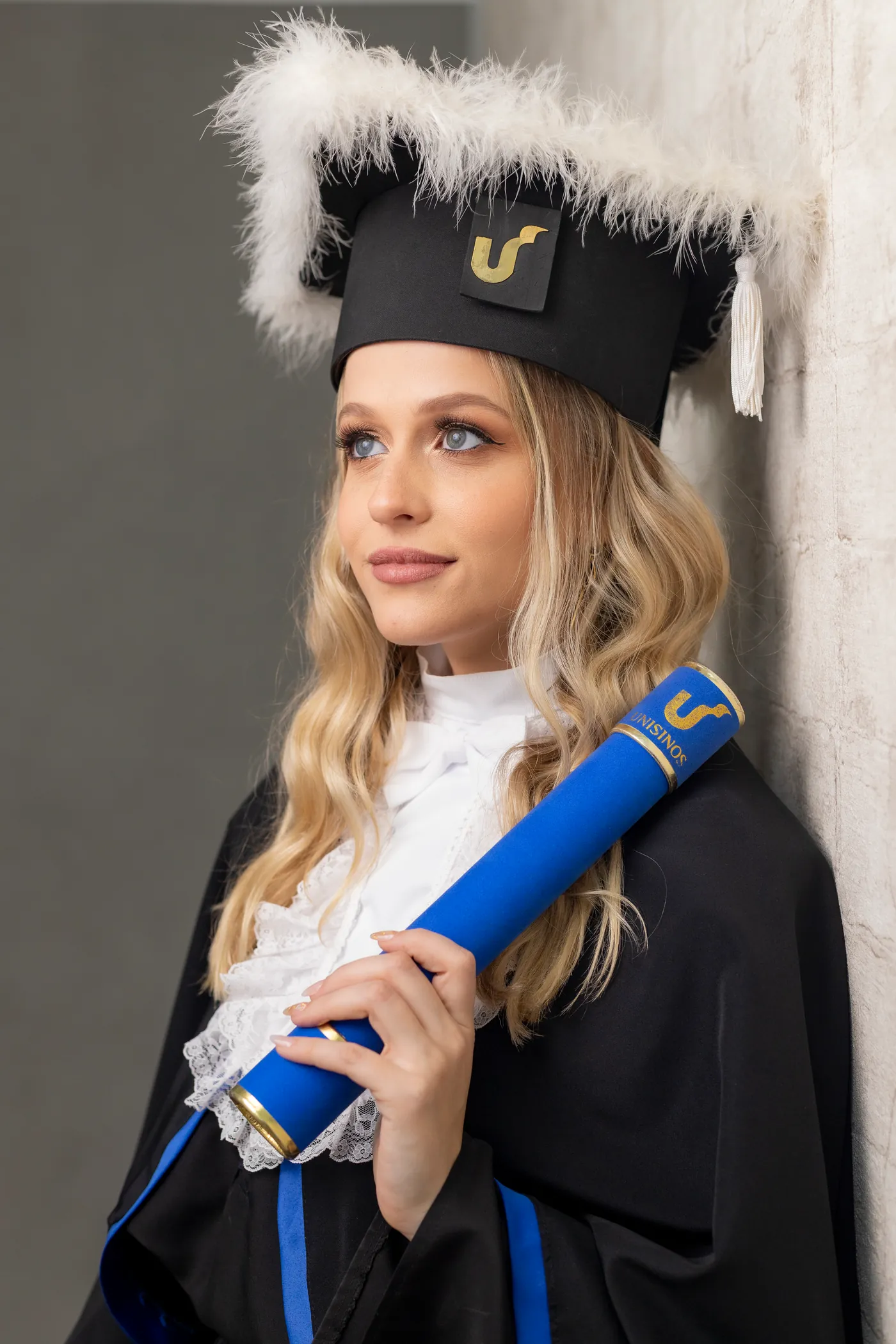 Nathália de beca e capelo, segurando o canudo azul da Unisinos e olhando para o lado